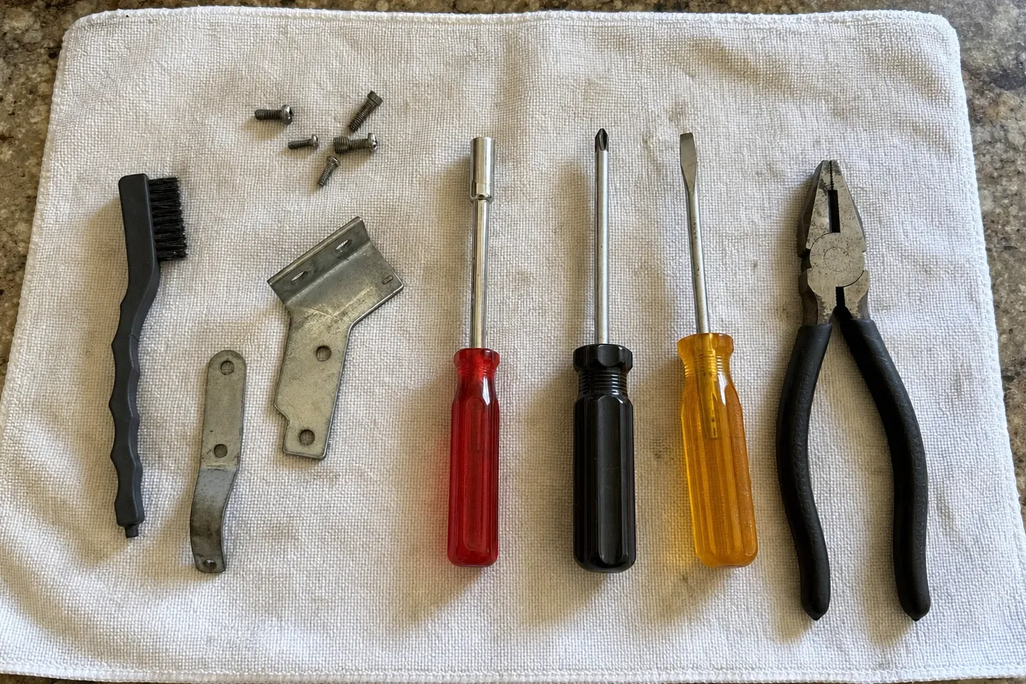 Wire brush, two steel brackets and six screws set out on a towel beside a nut driver, screwdrivers and pliers
