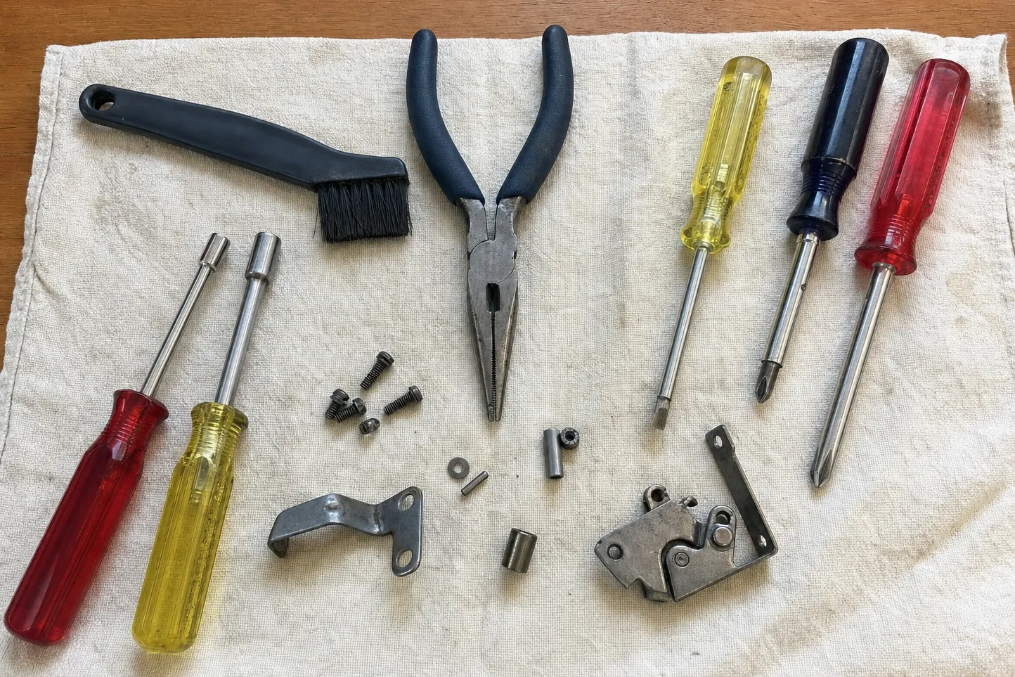 Latch assembly with its cam on an L-shaped mounting plate, laid out on a cream towel with a crank bracket, set screws, a roll pin, two spacers, a parts brush, pliers and five drivers