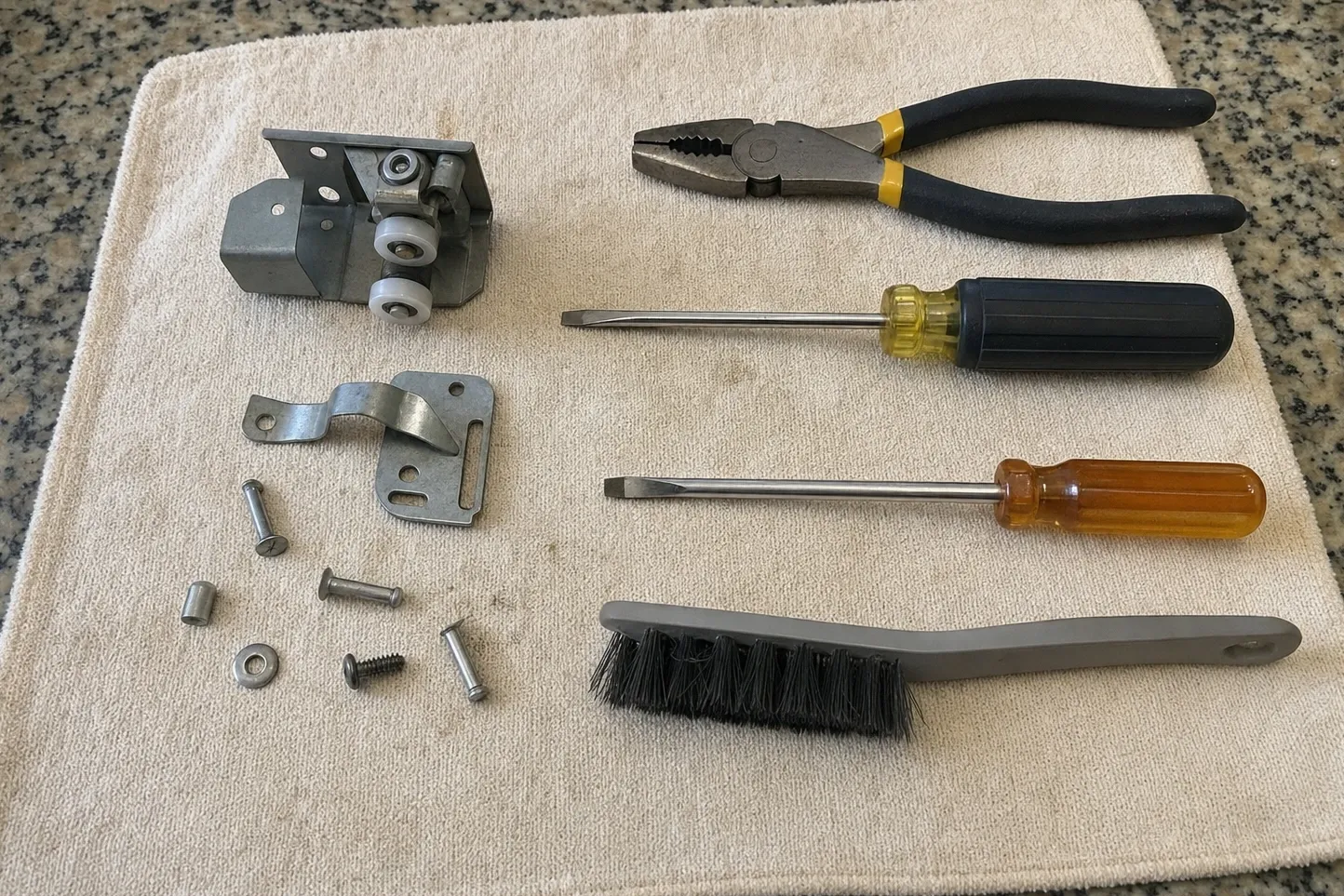 Roller carriage carrying two white nylon rollers on a folded steel mount, with a stepped strike plate, pins, a spacer and screws on a cream towel beside pliers, two drivers and a parts brush