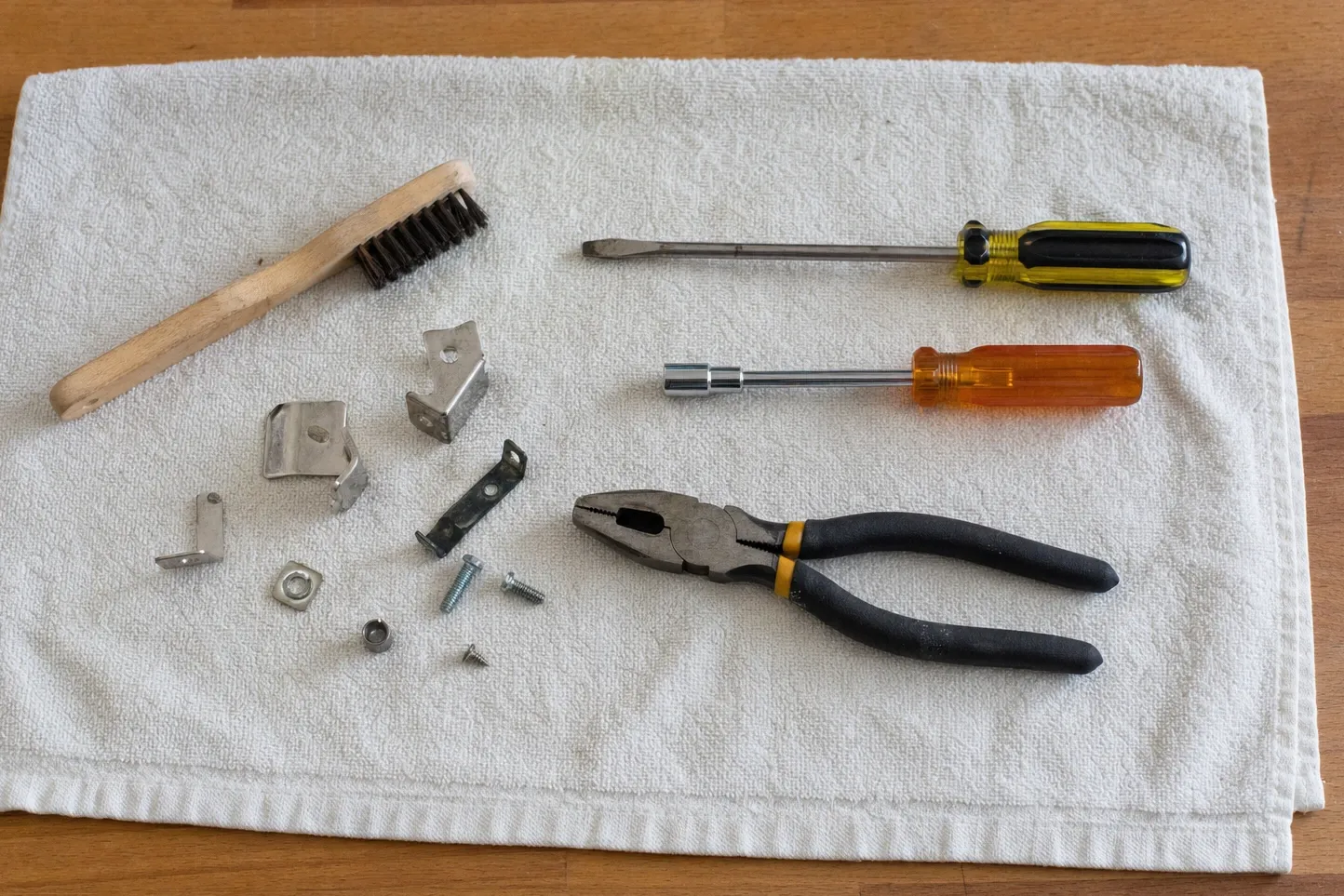 Four small stamped-steel support clips with a hex nut, a spacer and three screws on a white waffle towel, beside a wooden-handled wire brush, two drivers and lineman’s pliers