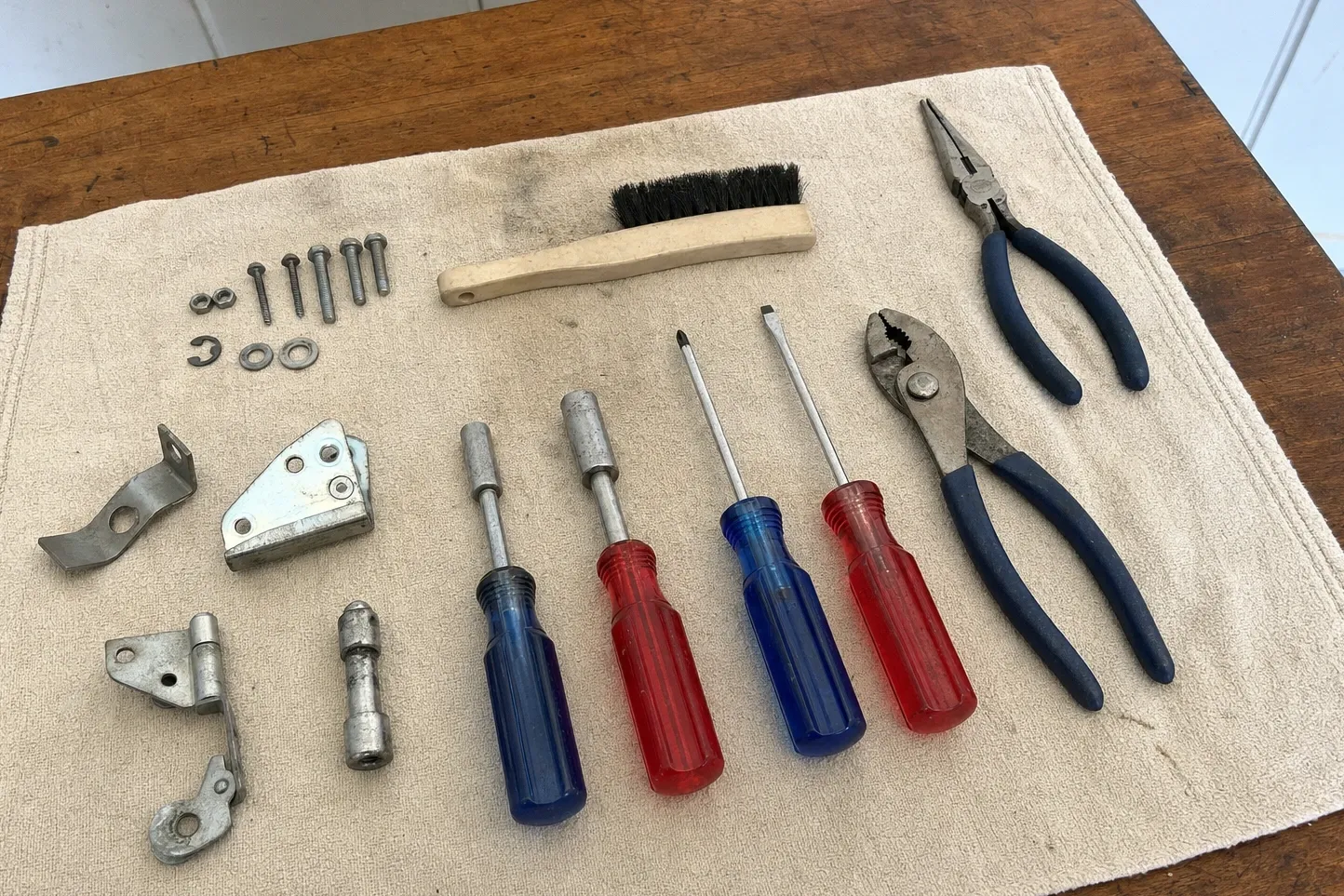 Stepped hinge arm with its roller boss and a triangular mounting plate laid out on a cream towel with a circlip, washers, machine screws, a steel spacer, four drivers, pliers and a wire brush